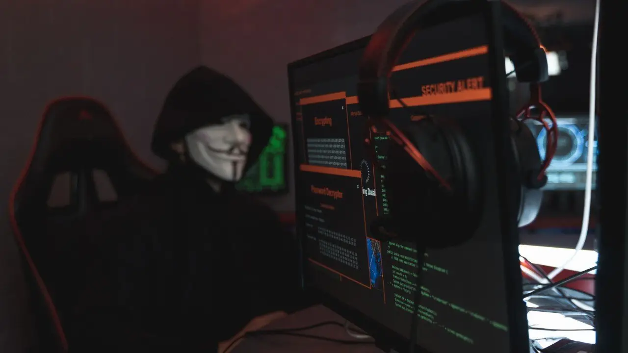 A hooded figure in a mask in front of screens showing a security alert