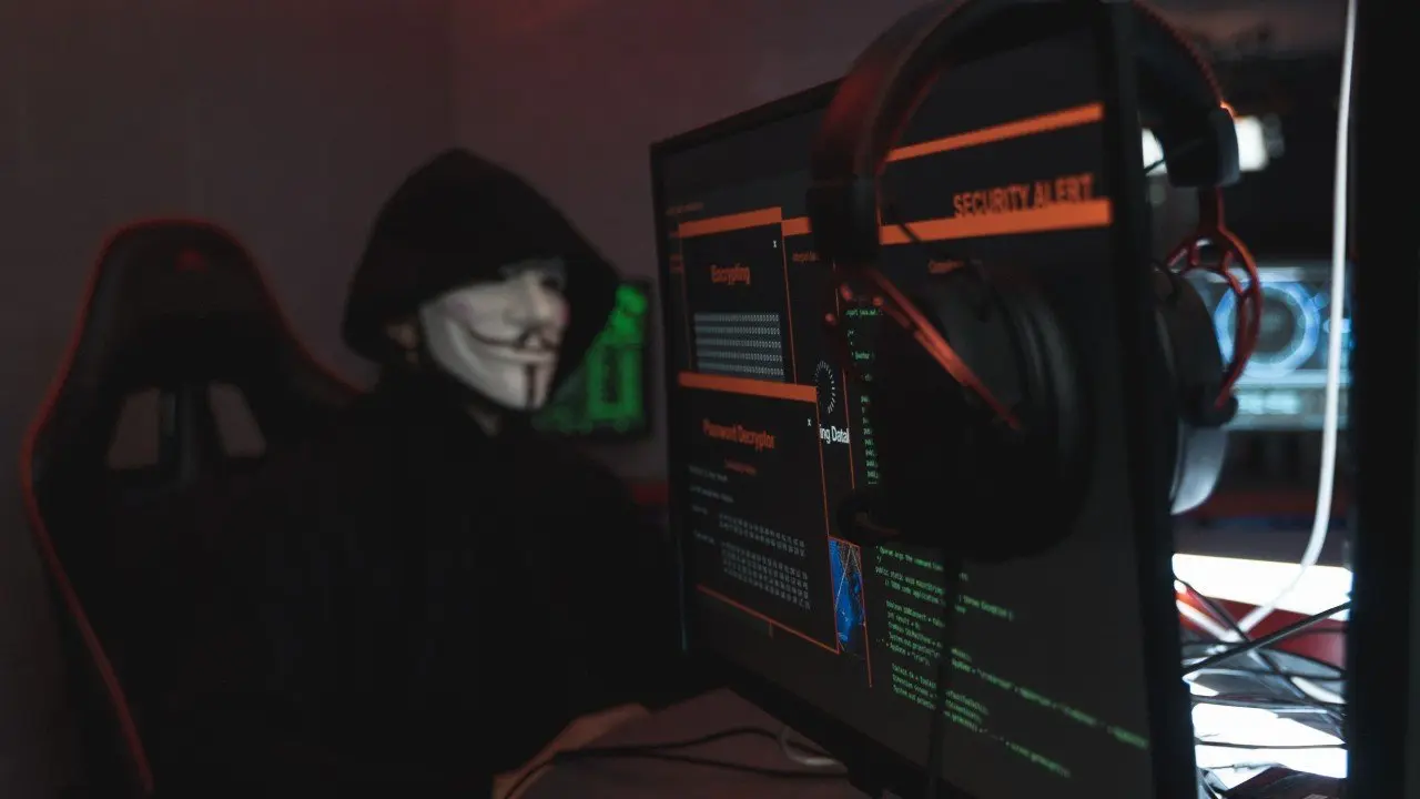 A hooded figure in a mask in front of screens showing a security alert